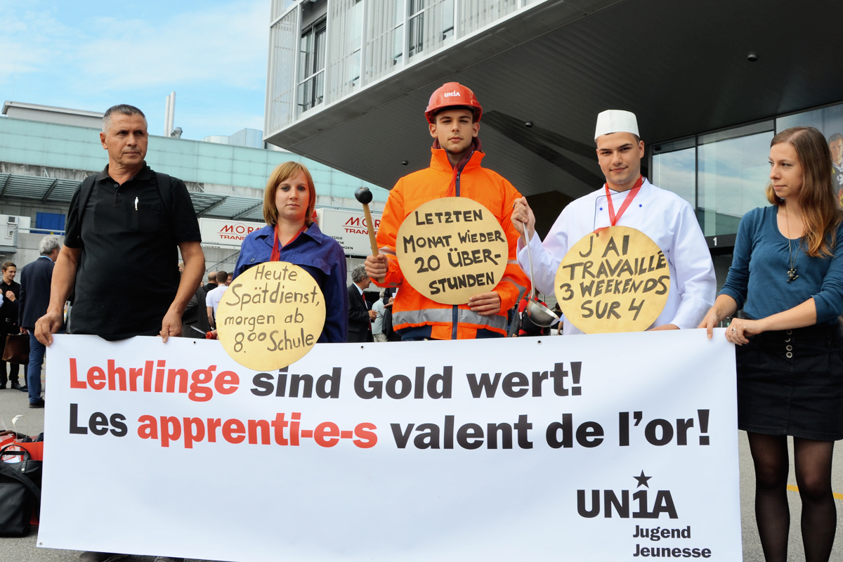 5 Gewerkschafter/innen mit Transparent und Plaketten: Lehrlinge sind Gold wert. Heute Spätdienst, morgen ab 8.00 Schule. Letzten Montat 20 Überstunden. J'ai travaillé 3 weekends sur 4.