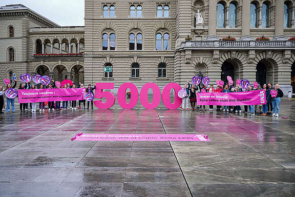 14 giugno, Giornata dello sciopero delle donne. Le donne sono in piedi davanti a un grande 5000 rosa e reggono uno striscione che recita: “Ancora incazzate: salari equi, NO al taglio della LPP!”