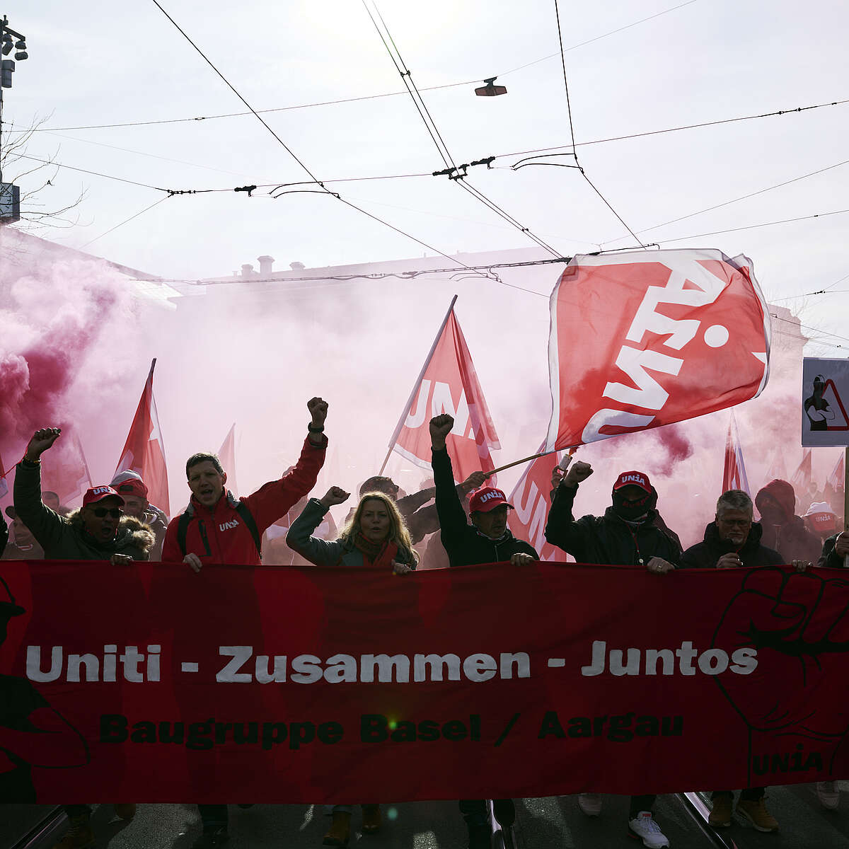 Persone manifestano con bandiere rosse e uno striscione «Uniti – Zusammen – Juntos» in una strada della città.