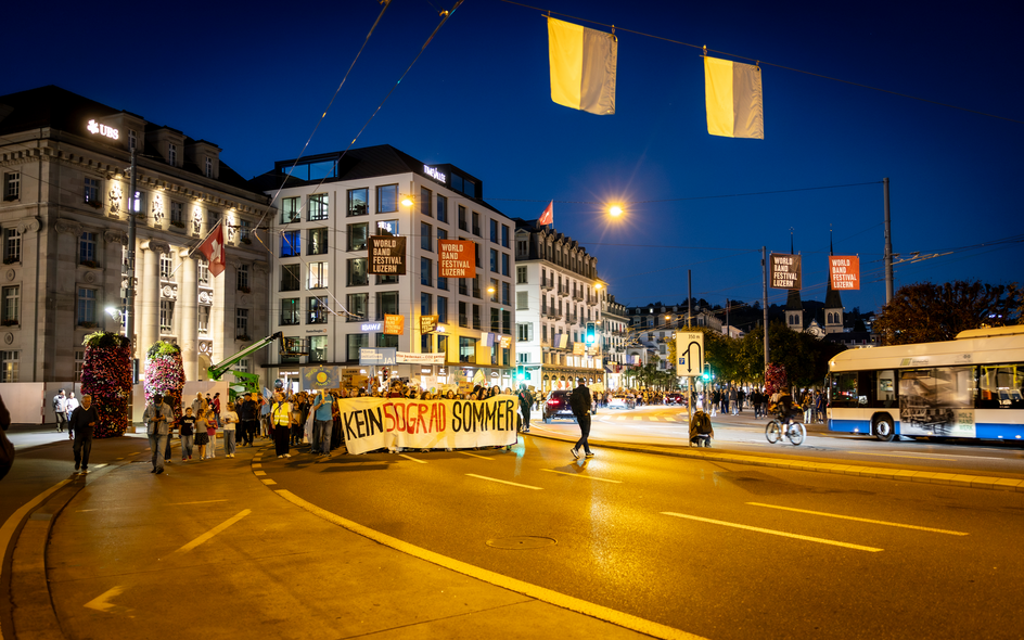 Un cortège au crépuscule dans une rue de Lucerne, entre façades éclairées et drapeaux.