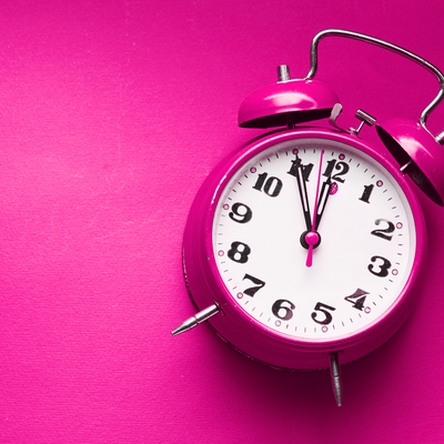 Ein pinker, altmodischer Wecker mit zwei Glocken liegt auf einem pinken Hintergrund. Die Zeiger stehen kurz vor zwölf. Der Wecker symbolisiert die Forderung nach mehr Zeit für Leben, Familie und Erholung – ein zentrales Anliegen zum 8. März, dem Tag der Frauenrechte.