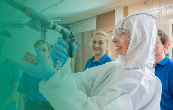 Eine Frau in Schutzanzug und Schutzbrille demonstriert den Umgang mit einem Reinigungsgerät vor einer Gruppe aufmerksam zuschauender Personen in blauer Arbeitskleidung.