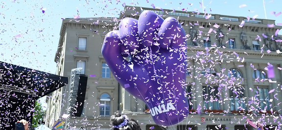 Ein grosser violetter Ballon in Form einer geballten Faust schwebt bei einer öffentlichen Demonstration vor einem Gebäude der Berner Innenstadt. Um die Faust herum fliegt violettes Konfetti durch die Luft. Auf der Faust steht Unia. Die Szene spielt sich bei Tageslicht unter blauem Himmel ab und vermittelt eine kämpferische und feierliche Stimmung.