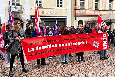 Un gruppo di manifestanti di diverse età tiene in piazza uno striscione rosso con scritta bianca e nera durante una manifestazione a Bellinzona, Svizzera