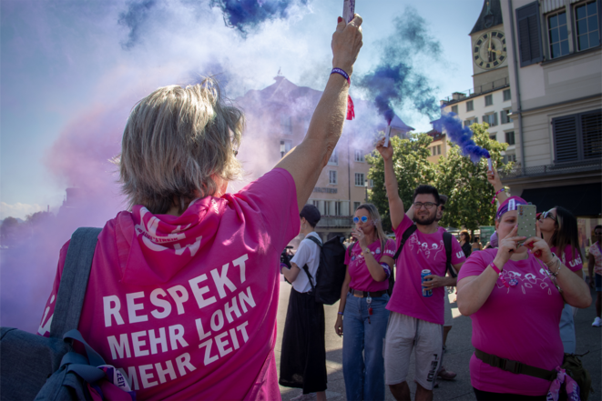 Das Bild zeigt eine Demonstration, bei der Menschen in pinken T-Shirts und mit feministischen Symbolen sichtbar sind. Eine Person im Vordergrund trägt ein T-Shirt mit der Aufschrift «Respekt, mehr Lohn, mehr Zeit.» Die Teilnehmenden halten bunte Rauchfackeln in die Höhe, die violetten Rauch erzeugen. Im Hintergrund sieht man eine städtische Umgebung mit Gebäuden und einer Turmuhr. Die Stimmung wirkt entschlossen und kämpferisch.