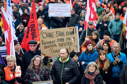 Nebeneinander stehende Menschen an einer Kundgebung halten Fahnen der Unia und anderer politischer Organisationen sowie ein Plakat mit den Worten: «Ohne Stahl kein Rückgrat – Steh zu unserer Industrie, Parmelin!»»