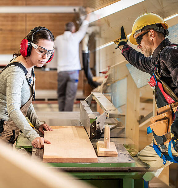 Una donna è al lavoro con una fresatrice da tavolo in una falegnameria; accanto a lei, un falegname con elmetto e cintura porta-attrezzi è impegnato nella costruzione in legno. 
