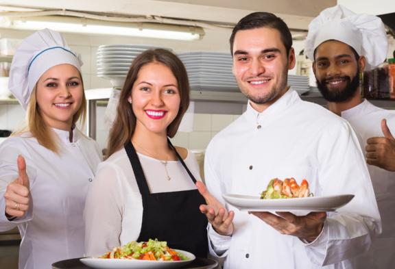 Une cuisinière, deux cuisiniers et une serveuse posent dans une cuisine de restaurant et sourient à la caméra. Deux tiennent des assiettes avec des plats, les autres montrent un pouce en l'air.