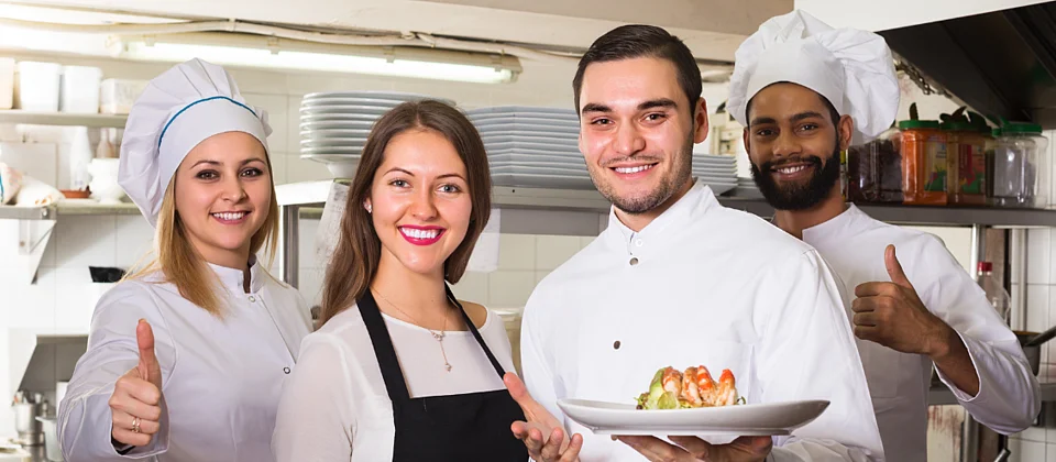 Una cuoca, due cuochi e una cameriera posano nella cucina di un ristorante e sorridono alla telecamera. Due di loro tengono in mano dei piatti con delle pietanze, gli altri mostrano il pollice alzato.