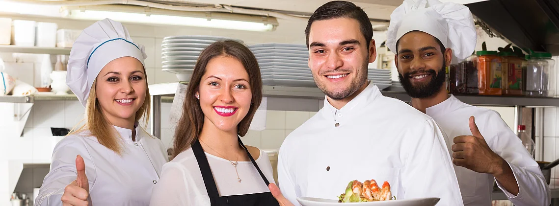  Eine Köchin, zwei Köche und eine Serviceangestellte posieren in einer Restaurantküche und lächeln in die Kamera. Zwei halten Teller mit Gerichten, die anderen zeigen Daumen hoch. 