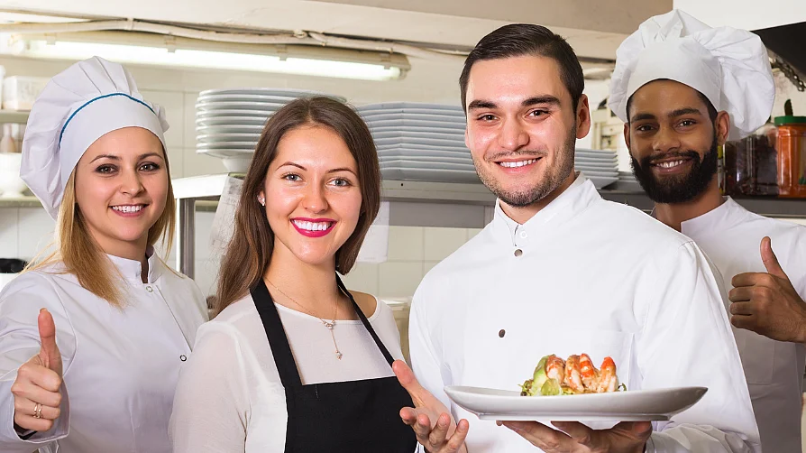 Eine Köchin, zwei Köche und eine Serviceangestellte posieren in einer Restaurantküche und lächeln in die Kamera. Zwei halten Teller mit Gerichten, die anderen zeigen Daumen hoch. 