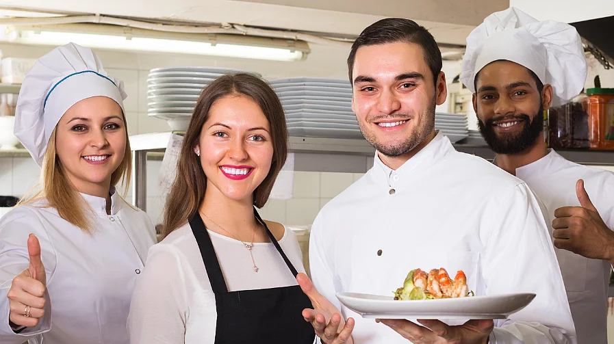 Una cuoca, due cuochi e una cameriera posano in una cucina di ristorante e sorridono alla fotocamera. Due tengono piatti con cibi, gli altri mostrano un pollice in su.