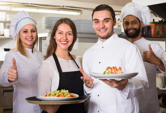 Una cuoca, due cuochi e una cameriera posano in una cucina di ristorante e sorridono alla fotocamera. Due tengono piatti con cibi, gli altri mostrano un pollice in su.