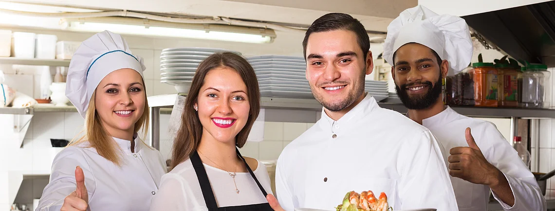 Una cuoca, due cuochi e una cameriera posano nella cucina di un ristorante e sorridono alla telecamera. Due di loro tengono in mano dei piatti con delle pietanze, gli altri mostrano il pollice alzato.