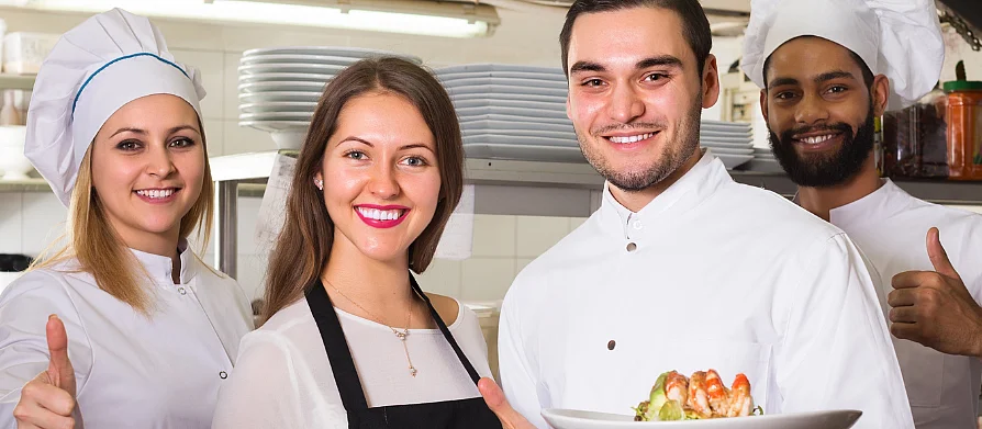 Eine Köchin, zwei Köche und eine Serviceangestellte posieren in einer Restaurantküche und lächeln in die Kamera. Zwei halten Teller mit Gerichten, die anderen zeigen Daumen hoch. 