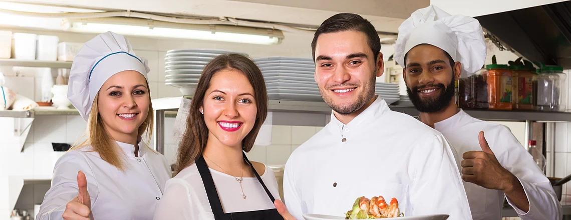 Une cuisinière, deux cuisiniers et une serveuse posent dans la cuisine d'un restaurant et sourient à la caméra. Deux d'entre eux tiennent des assiettes avec des plats, les autres lèvent le pouce.