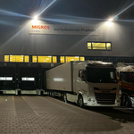 Un centre logistique de Migros Online à Pratteln (BL) la nuit. Des camions sont aux quais de chargement, le bâtiment est éclairé.