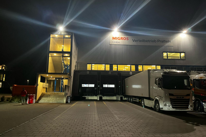 Un centre logistique de Migros Online à Pratteln (BL) la nuit. Des camions sont aux quais de chargement, le bâtiment est éclairé.