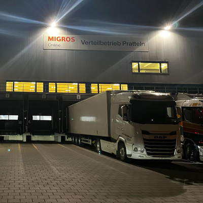 Ein Logistikzentrum von Migros Online in Pratteln (BL) bei Nacht. LKWs stehen an den Verladerampen, das Gebäude ist beleuchtet.