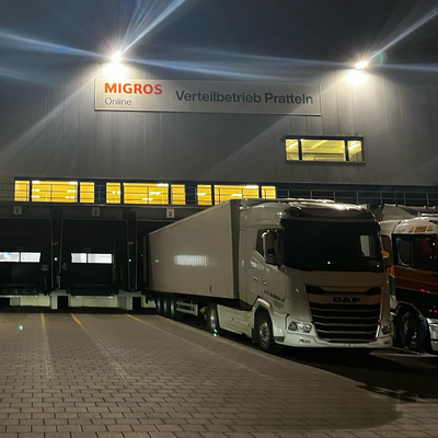 Un centre logistique de Migros Online à Pratteln (BL) la nuit. Des camions sont aux quais de chargement, le bâtiment est éclairé.