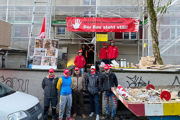 Geprellte Bauarbeiter vor der Baustelle und einem Transparent: Wir kämpfen für unsere Gesundheit! Der Bau steht still!