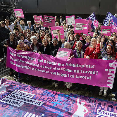 Ca. 50 Frauen stehen hinter einem Transparent auf dem in drei Sprachen «Stopp Belästigung und Gewalt am Arbeitsplatz» steht. Sie halten violette und rote Fahnen sowie pinke Schilder mit weiteren Botschaften.