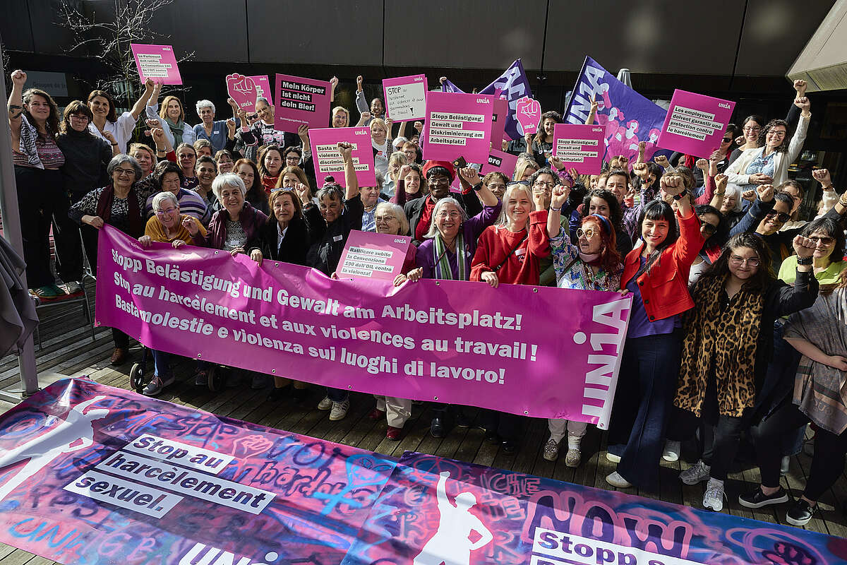 Ca. 50 Frauen stehen hinter einem Transparent auf dem in drei Sprachen «Stopp Belästigung und Gewalt am Arbeitsplatz» steht. Sie halten violette und rote Fahnen sowie pinke Schilder mit weiteren Botschaften.