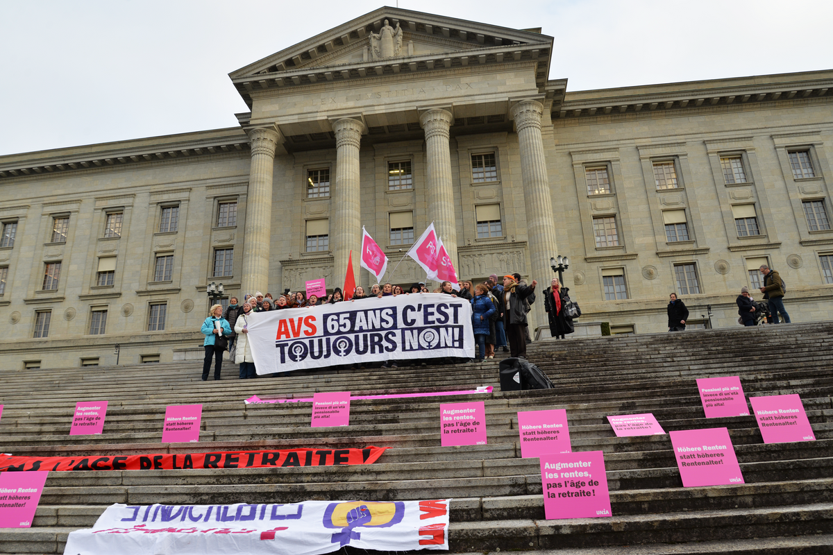 Un groupe de personnes se tient sur les escaliers devant le Tribunal fédéral à Lausanne et brandit des bannières et des affiches roses. Sur la bannière centrale, on peut lire : « Augmenter les rentes, pas l’âge de la retraite ! » D'autres panneaux et bannières en allemand et en français demandent de meilleures conditions de vie pour les femmes retraitées et s'opposent à l'augmentation de l'âge de la retraite des femmes à 65 ans. Certaines participantes brandissent des drapeaux syndicaux, d'autres des pancartes plus petites. La scène montre une action de manifestation pacifique avec un message clair. 