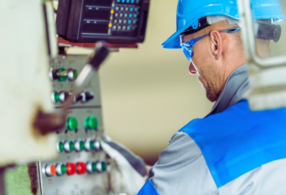 Un travailleur avec un casque et des lunettes de protection utilise un panneau de commande avec différents boutons et réglages.