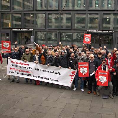 Eine Gruppe von rund 50 Personen ist in einer urbanen Umgebung zu sehen. Sie halten ein Transparent und einige Hohlkammerplakate. Auf dem Transparent steht «Maler-Gipser: Für unseren GAV – für unsere Zukunft.» sowie «Pittoria-gessattorie: Per il nostro CCL, per il nostro futuro.»