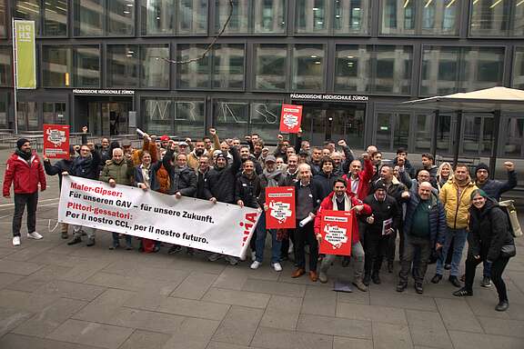 Eine Gruppe von rund 50 Personen ist in einer urbanen Umgebung zu sehen. Sie halten ein Transparent und einige Hohlkammerplakate. Auf dem Transparent steht «Maler-Gipser: Für unseren GAV – für unsere Zukunft.» sowie «Pittoria-gessattorie: Per il nostro CCL, per il nostro futuro.»