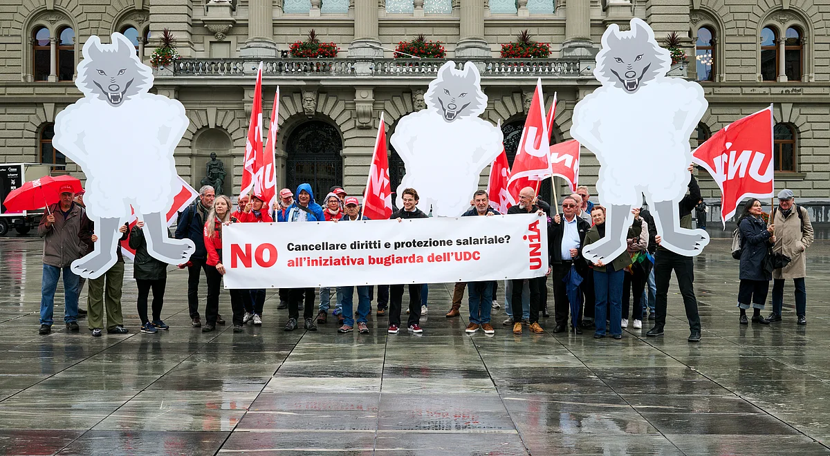 I manifestanti davanti al Palazzo federale tengono uno striscione con la scritta «NO all'iniziativa bugiarda dell'UDC» e protestano con bandiere Unia e grandi figure di lupi travestiti da agnelli contro lo smantellamento dei diritti e della tutela salariale.