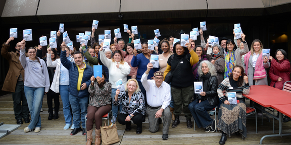 Eine grosse, vielfältige Gruppe von Menschen hält Broschüren mit dem Titel «Care Manifest - für gute Pflege und Betreuung» in die Höhe und lächelt in die Kamera.