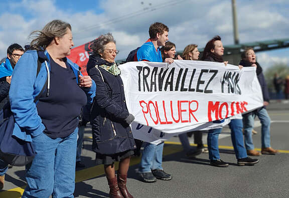Un groupe de personnes manifeste dans la rue en tenant une banderole sur laquelle est écrit: «Travaillez moins, polluez moins»