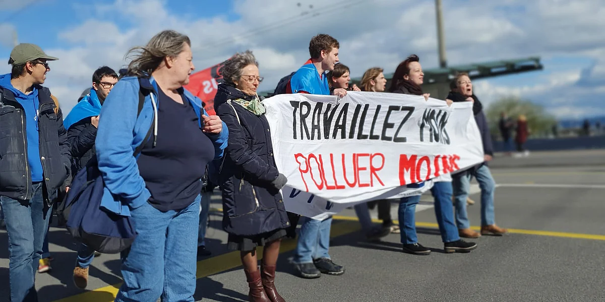 Un groupe de personnes manifeste dans la rue en tenant une banderole sur laquelle est écrit: «Travaillez moins, polluez moins»