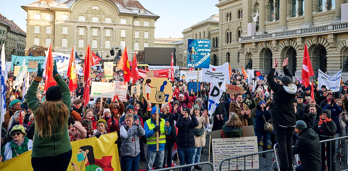 Il personale curante manifesta con striscioni, bandiere e cartelli sulla piazza Federale a Berna, in primo piano sono visibili messaggi sulle condizioni di lavoro e sull'uguaglianza dei sessi.