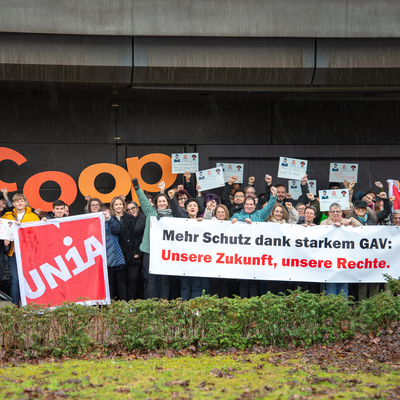 Eine Gruppe von 50 Menschen halten ein Transparent, Fahnen, Kartonschilder und grosse ausgestanzte Buchstaben, welche dem Firmenlogo von Coop nachempfunden sind. Sie strecken ihre Faust in die Luft und erwecken einen kämpferischen gewerschaftlichen Eindruck. Auf dem Transparent steht: «Mehr Schutz dank starkem GAV: Unsere Zukunft, unsere Rechte.»