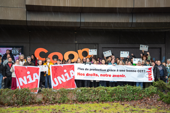 Un groupe de 50 personnes brandissent une banderole, des drapeaux, des pancartes en carton et de grandes lettres découpées inspirées du logo Coop. Ils lèvent le poing en l’air et affichent l’image d’un syndicat combatif. Sur la banderole, on peut lire: «Une meilleure protection grâce à une CCT forte: notre avenir, nos droits.»