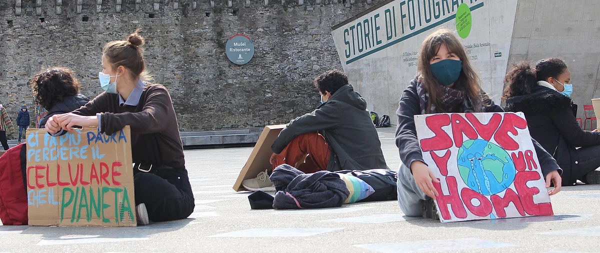 Giovani seduti a terra indossano mascherine e mostrano cartelli di protesta: uno con la scritta “SAVE YOUR HOME” e un disegno del pianeta che piange, l’altro con “Ci fa più paura perdere il cellulare che il pianeta”.