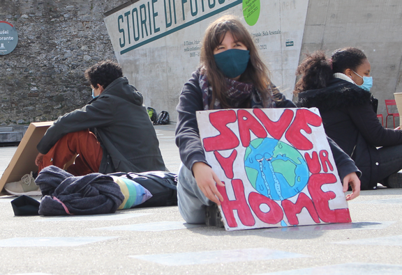 Giovani seduti a terra indossano mascherine e mostrano cartelli di protesta: uno con la scritta “SAVE YOUR HOME” e un disegno del pianeta che piange, l’altro con “Ci fa più paura perdere il cellulare che il pianeta”.