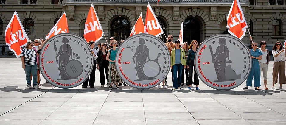 Unia-Aktivist:innen vor dem Bundeshaus in Bern halten drei übergrosse Münzen mit Helvetia und der Aufschrift «Stopp Lohnsenkungen per Gesetz» in den Landessprachen.