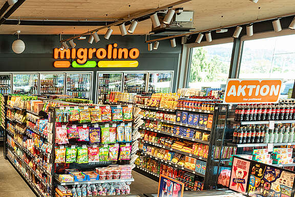 Innenansicht eines Verkaufsgeschäfts. Regale mit verschiedenen Kiosk-Produkten, einigen Lebensmitteln, einem Kühlgestell mit Getränken und einer Kassentheke sind zu sehen.