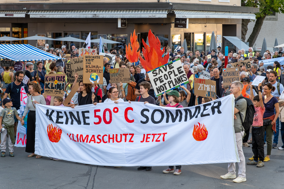 Des manifestant-e-s avec des drapeaux rouges derrière une banderole blanche portant l’inscription «Keine 50°C Sommer».
