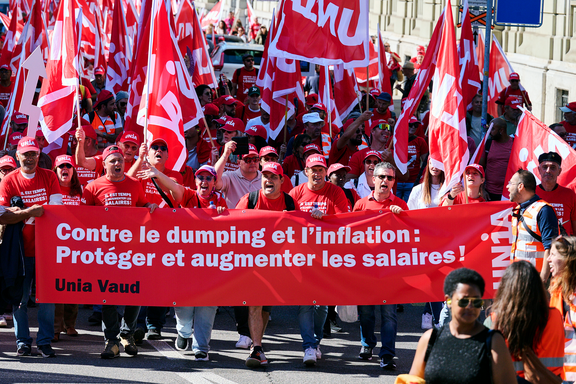 Manifestation syndicale organisée par Unia Vaud: une foule de personnes marche en brandissant des drapeaux rouges et une banderole avec l’inscription 'Contre le dumping et l’inflation : Protéger et augmenter les salaires !' Les participant-e-s portent des casquettes et des t-shirts aux couleurs du syndicat.