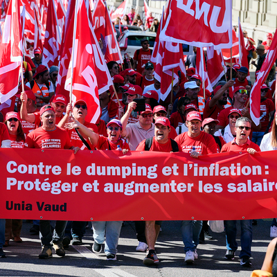 Manifestation syndicale organisée par Unia Vaud: une foule de personnes marche en brandissant des drapeaux rouges et une banderole avec l’inscription 'Contre le dumping et l’inflation : Protéger et augmenter les salaires !' Les participant-e-s portent des casquettes et des t-shirts aux couleurs du syndicat.