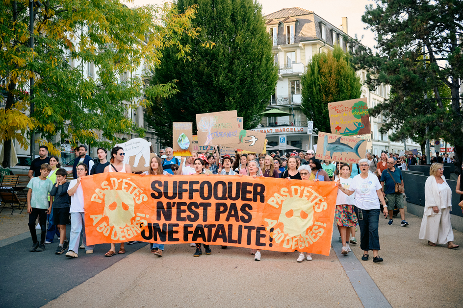 Des manifestant-e-s de tous âges derrière une banderole orange, dans une rue bordée d’arbres.