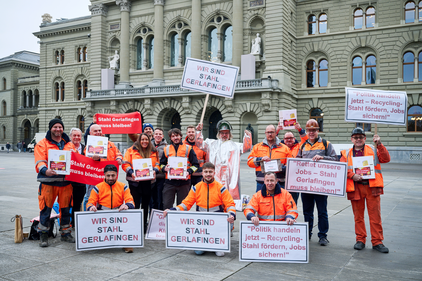 Rund 20 Arbeiter:innen der Stahl Gerlafingen in orangen Warnjacken und zum Teil auch -hosen vor dem Bundeshaus in Bern. Sie tragen Schilder mit Aufschriften wie: Wir sind Stahl Gerlafingen. Oder: Politik handeln jetzt - Recyclinkg-Stahl fördern, Jobs sichern!