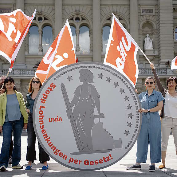 Aktion vor dem Bundesrat in Bern: Mehrere Menschen - darunter die Präsidentin der Gewerkschaft Unia Vania Alleva - stehen mit Unia-Fahnen rund um 2-Meter-hohen stilisierten 5-Franken-Münzen aus Karton mit der Aufschrift «Stopp Lohnsenkungen per Gesetz!»