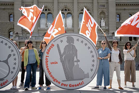 Aktion vor dem Bundesrat in Bern: Mehrere Menschen - darunter die Präsidentin der Gewerkschaft Unia Vania Alleva - stehen mit Unia-Fahnen rund um 2-Meter-hohen stilisierten 5-Franken-Münzen aus Karton mit der Aufschrift «Stopp Lohnsenkungen per Gesetz!»