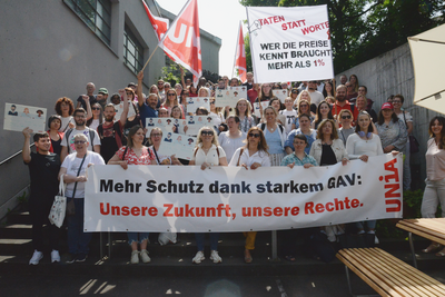  150 Personen stehen auf einer Treppe. Sie halten Kartonschilder und Fahnen in den Händen. Die vorderste Reihe hält ein Transparent, auf dem «Mehr Schutz dank starkem GAV: Unsere Zukunft, unsere Rechte.» steht.